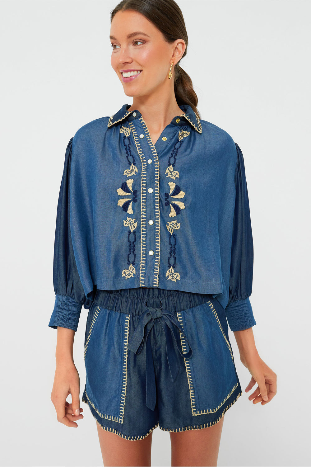 A woman wears a Carolina K. embroidered denim Elisa Shirt with buttons, paired with high-waisted drawstring shorts featuring contrast stitching.