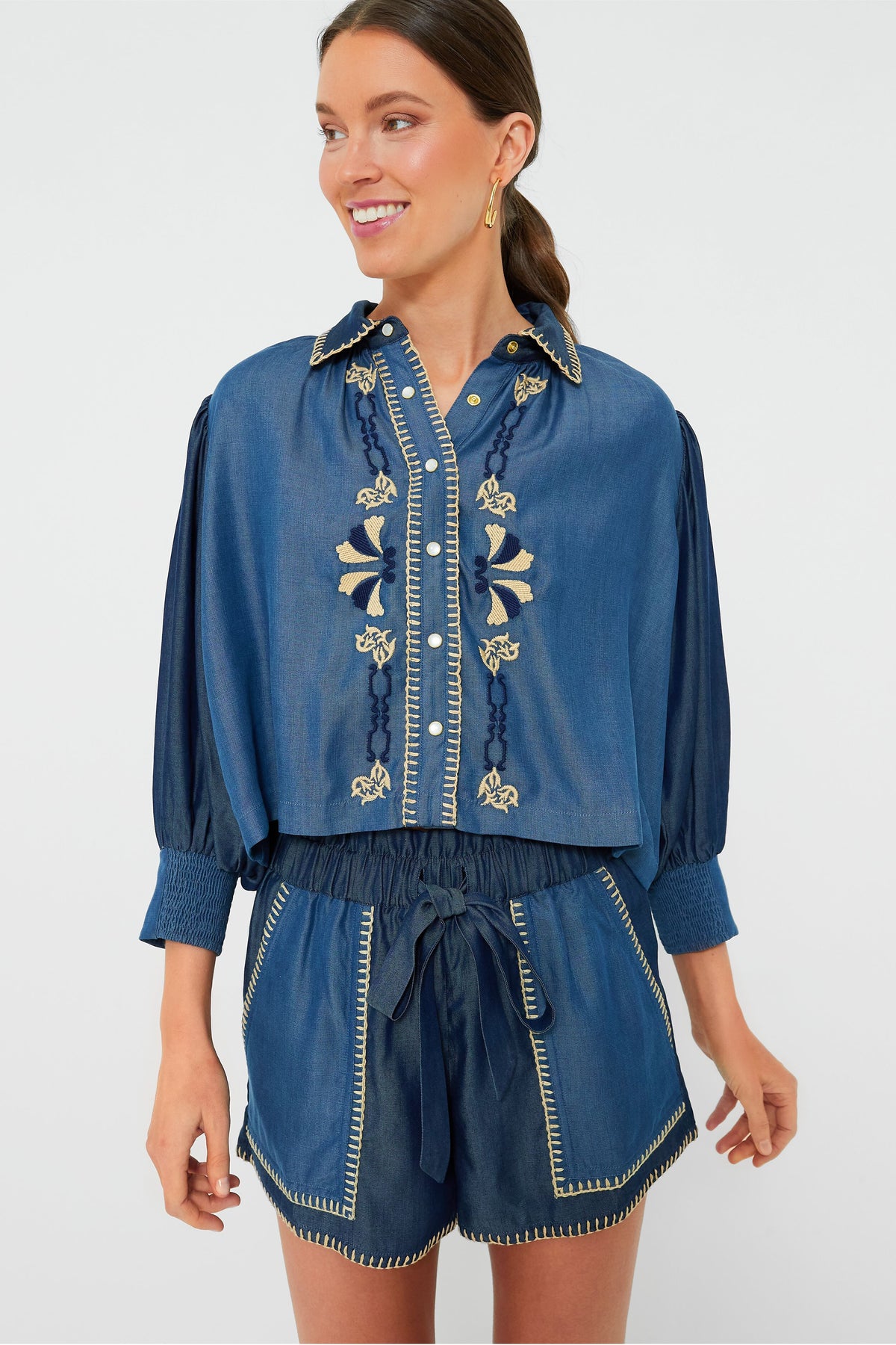 A woman wears a Carolina K. embroidered denim Elisa Shirt with buttons, paired with high-waisted drawstring shorts featuring contrast stitching.