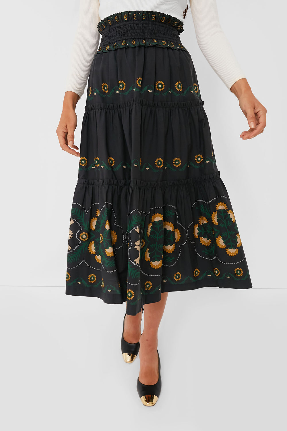 A person wearing a Sea New York high rise midi skirt with green and yellow floral embroidery and a smocked waistband, paired with a white ribbed top and black shoes featuring gold toe caps.