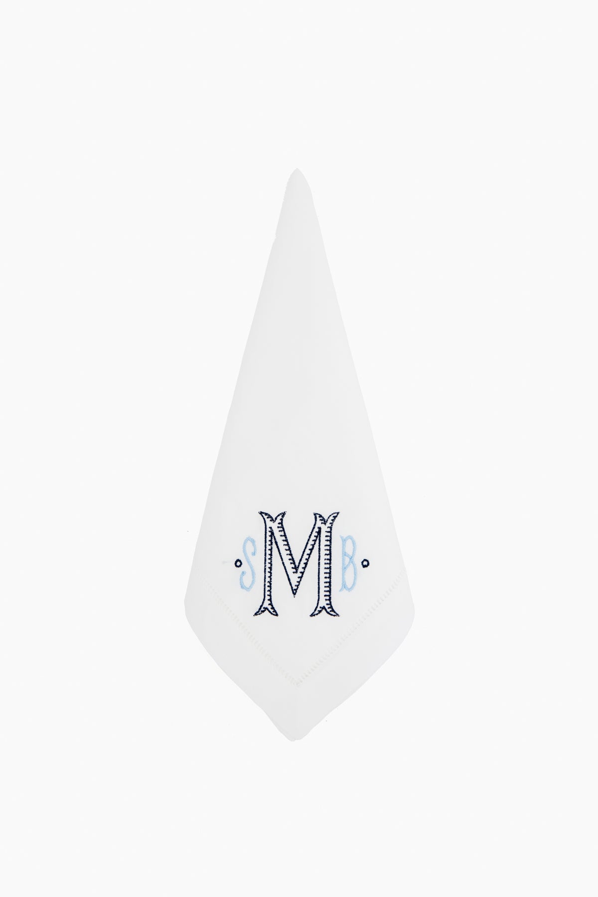 A white linen-cotton napkin by Caton's Wharf is folded into a triangle and features a decorative embroidered letter M with blue accents in the center, making it an ideal personalized gift.