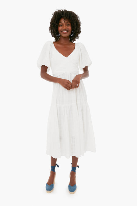 A woman smiles with her hands clasped in front, wearing a Hyacinth House white puff-sleeve midi dress and blue espadrille wedge sandals.
