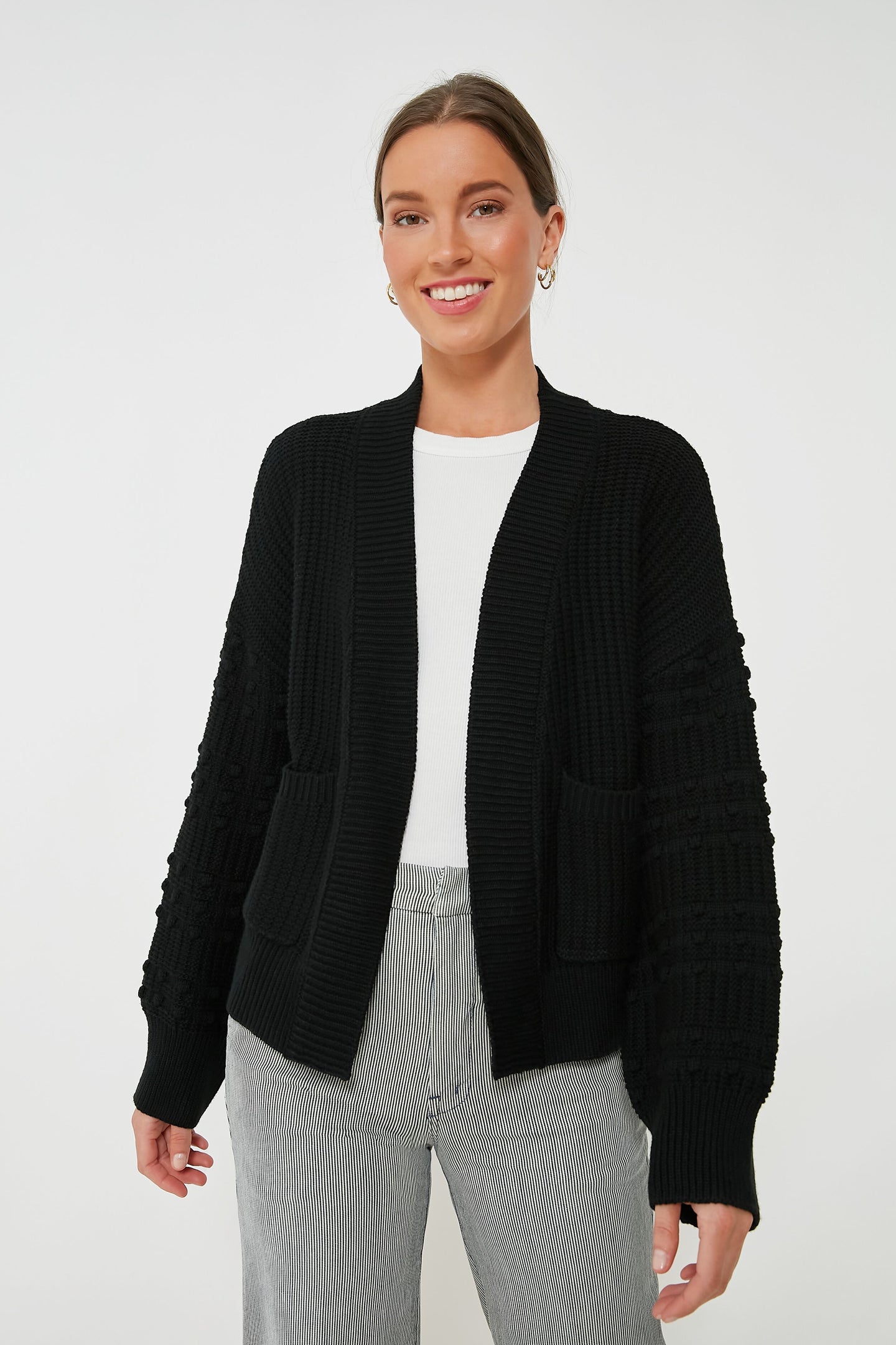 Woman standing and smiling, wearing a Pomander Place Black Bobble Cardigan with pom accents over a white top and gray pinstriped pants.