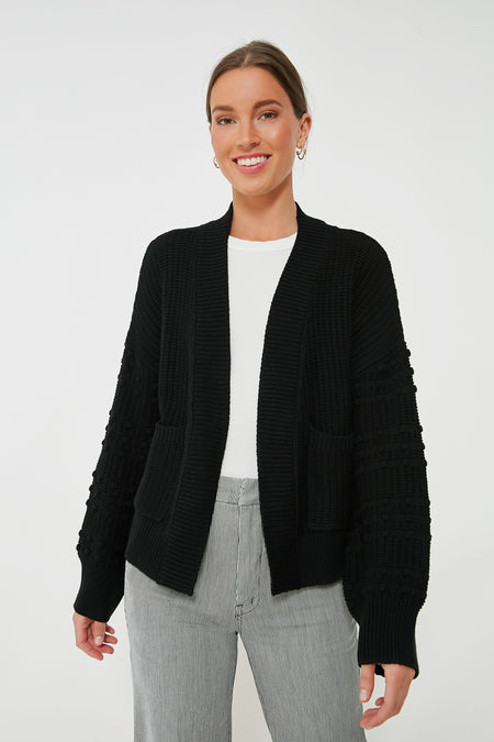 Woman standing and smiling, wearing a Pomander Place Black Bobble Cardigan with pom accents over a white top and gray pinstriped pants.