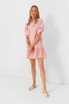 Woman in a pink Pomander Place Beatrix mini dress with ruffled sleeves and white slide sandals, smiling with hands loosely clasped.