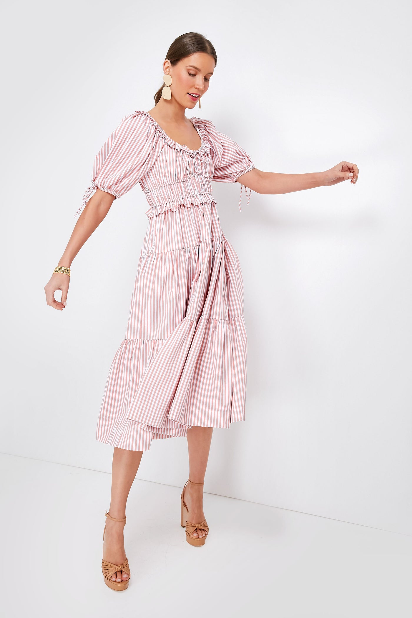 A woman wears the Pink Striped Brielle Dress by En Saison, a feminine spring dress with puff sleeves and ruffled details, paired with tan high-heeled sandals and large round earrings.
