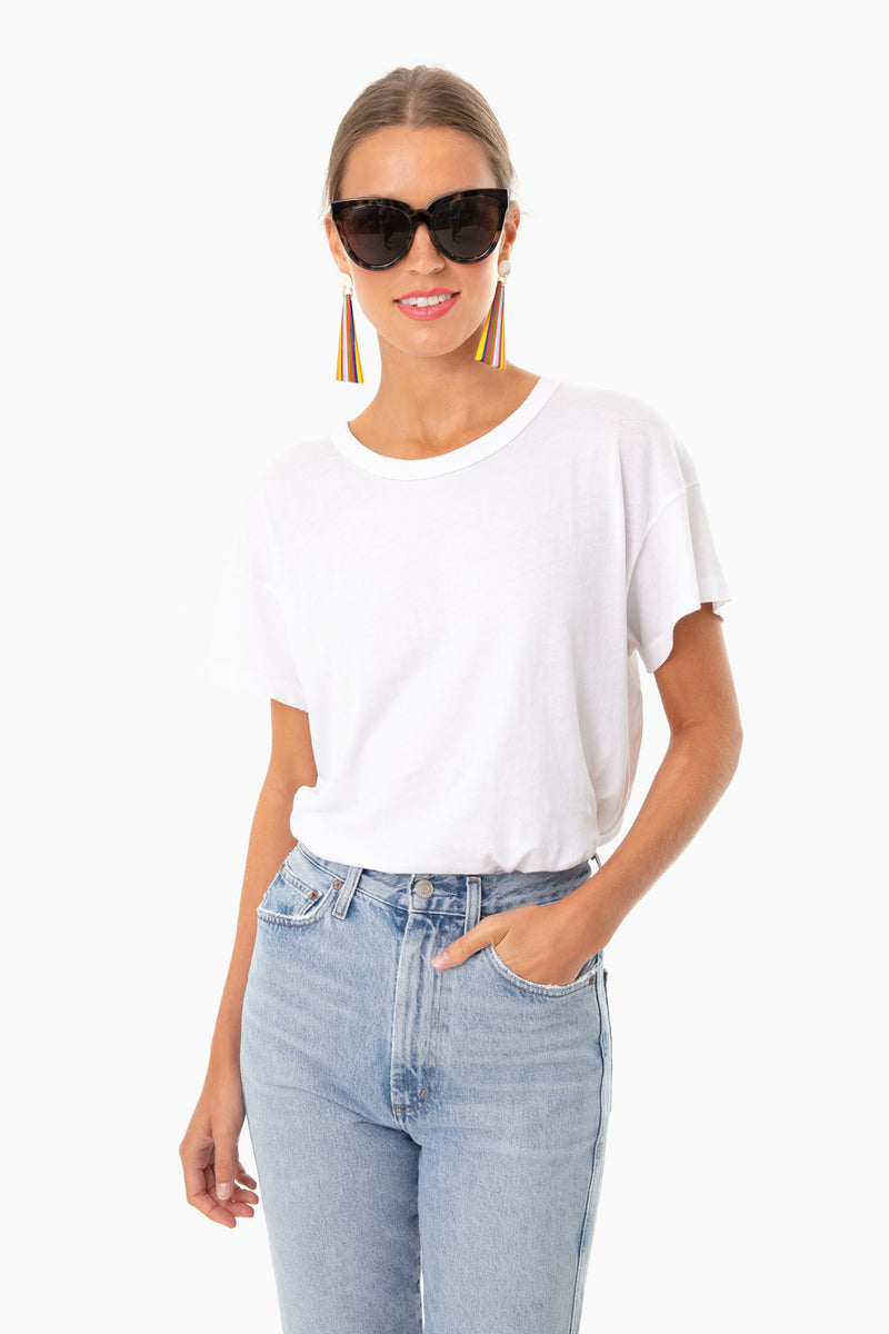 A woman wears THE GREAT. 100% cotton white boxy crew tee, light blue jeans, large black sunglasses, and colorful tassel earrings.