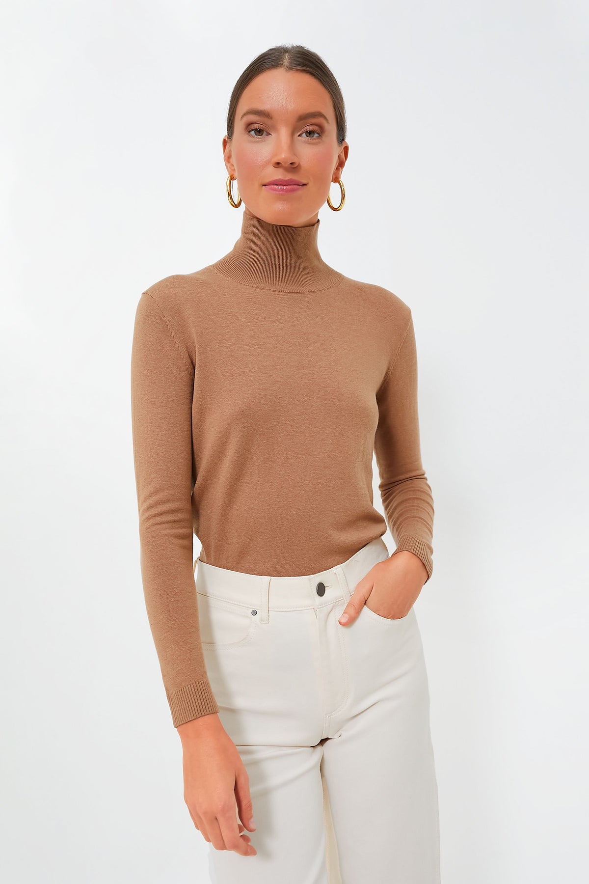 A woman in a brown silk-wool turtleneck sweater and white pants from Weekend Max Mara stands with one hand in her pocket, embodying casual elegance.