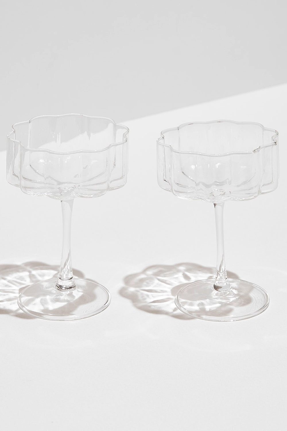 Two empty, modern FAZEEK glassware clear wave coupes with scalloped rims are placed side by side on a white surface, casting elegant shadows.