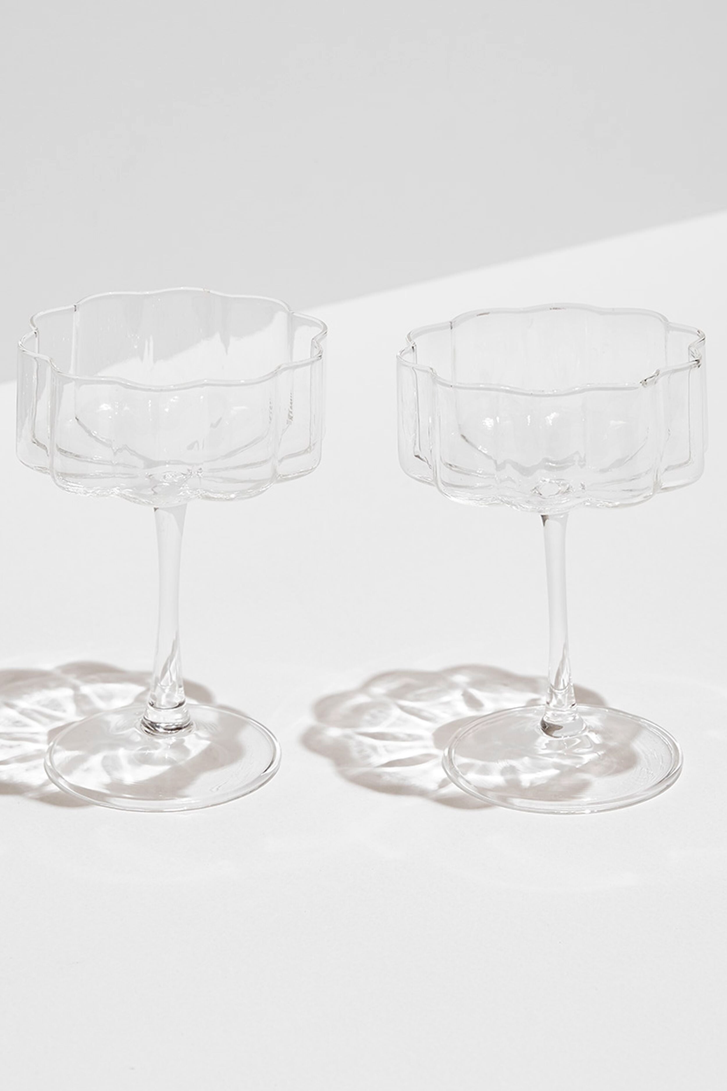 Two empty, modern FAZEEK glassware clear wave coupes with scalloped rims are placed side by side on a white surface, casting elegant shadows.