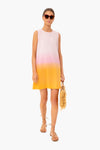A woman in sunglasses wears a KULE ombré sunset dress in white, pink, and yellow, holding a straw bag and wearing flat sandals.