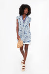 A woman in a blue and white floral Poupette St Barth dress holds a woven bag and wears sandals.