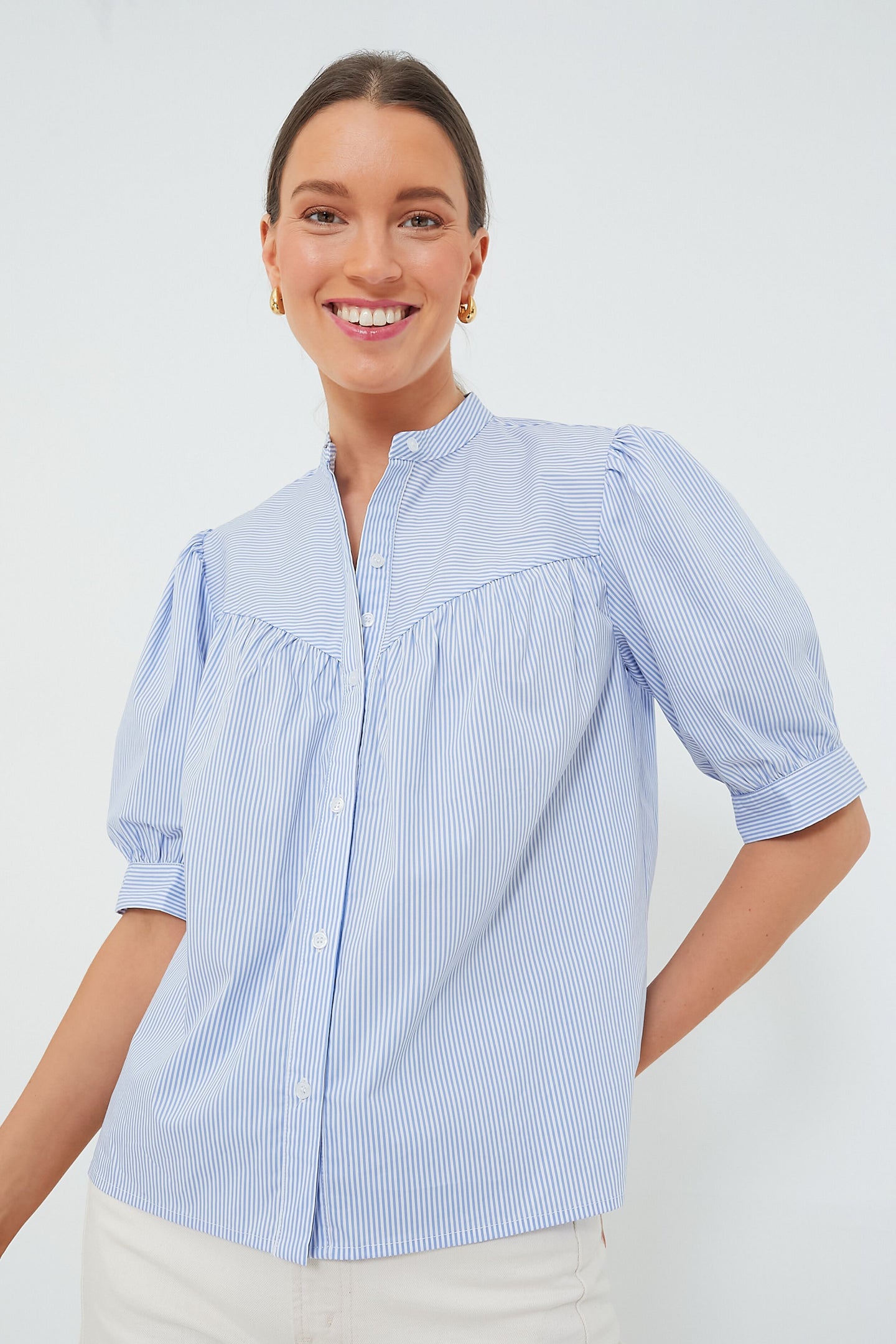 A woman smiles in a light blue and white vertical striped No Gape shirt from The Shirt by Rochelle Behrens, styled with puffed short sleeves and gold hoop earrings.