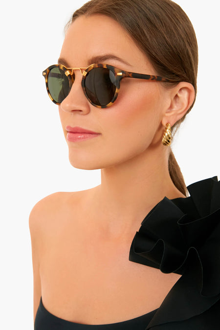 A woman wears KREWE du optic polarized, handcrafted acetate tortoiseshell sunglasses, gold twisted hoop earrings, and a black off-shoulder top with a large ruffle detail.