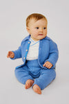 A baby sits barefoot on the floor in a light blue merino wool Oso & Me baby jacket and pants over a white shirt—ideal for parents wanting durable baby clothes.