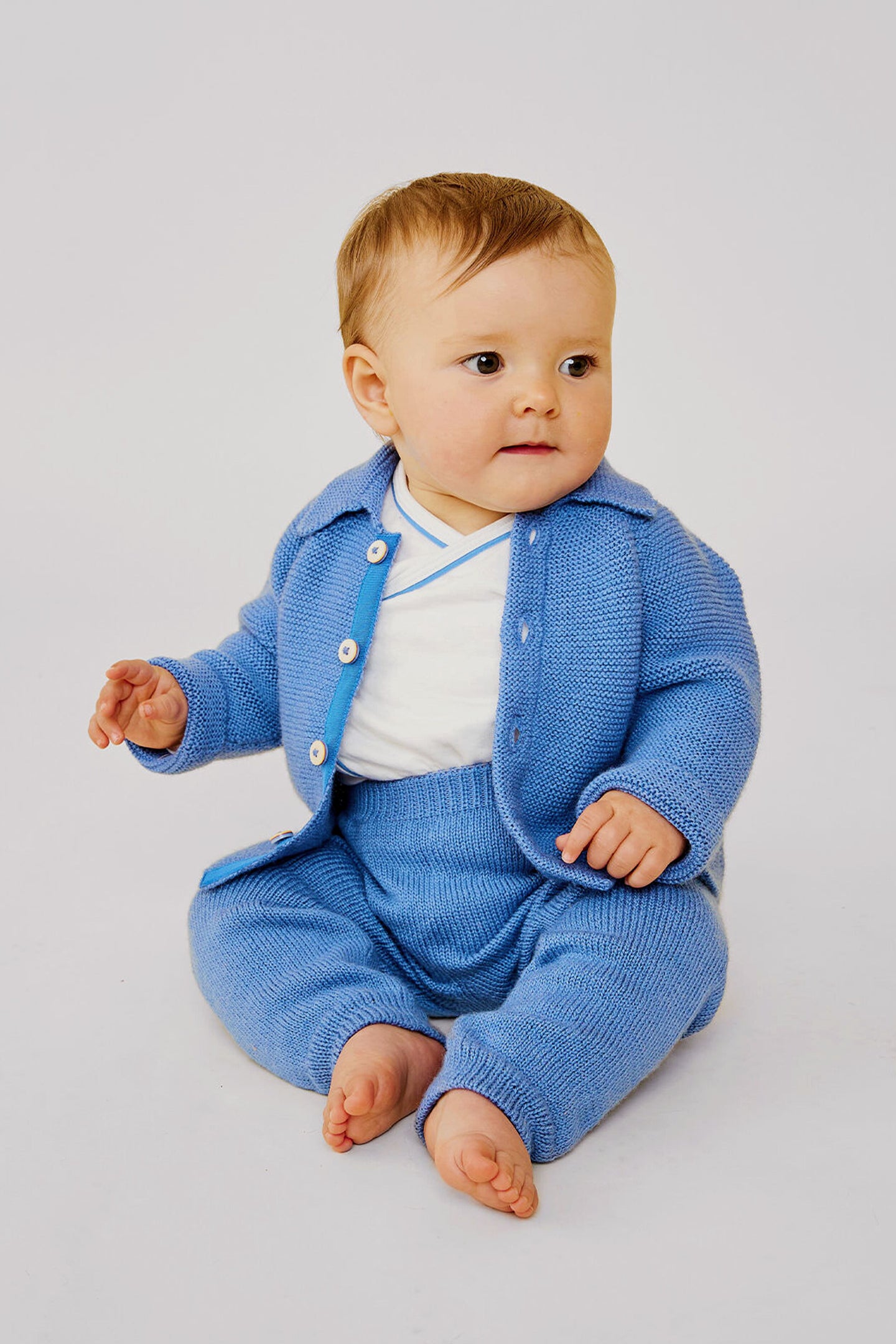 A baby sits barefoot on the floor in a light blue merino wool Oso & Me baby jacket and pants over a white shirt—ideal for parents wanting durable baby clothes.