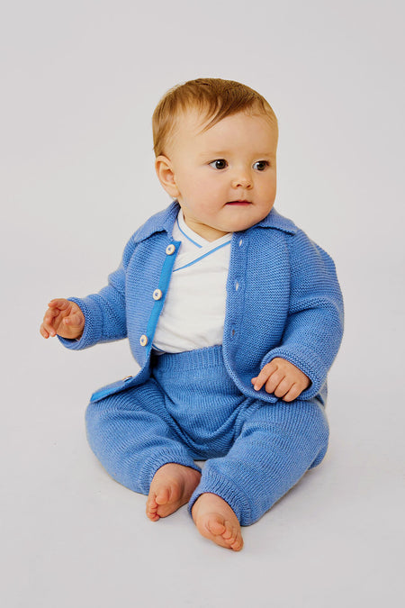 A baby sits barefoot on the floor in a light blue merino wool Oso & Me baby jacket and pants over a white shirt—ideal for parents wanting durable baby clothes.
