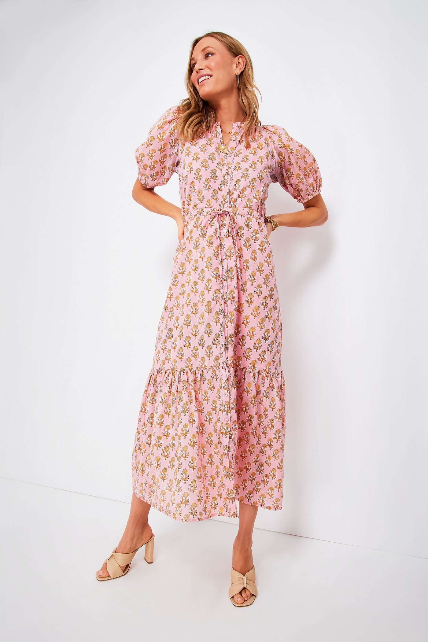 A woman wears a pink, floral, puff-sleeve organic cotton midi dress with a tie waist from the India Collection by Love Tomas by Emerson Fry, paired with beige heeled sandals.