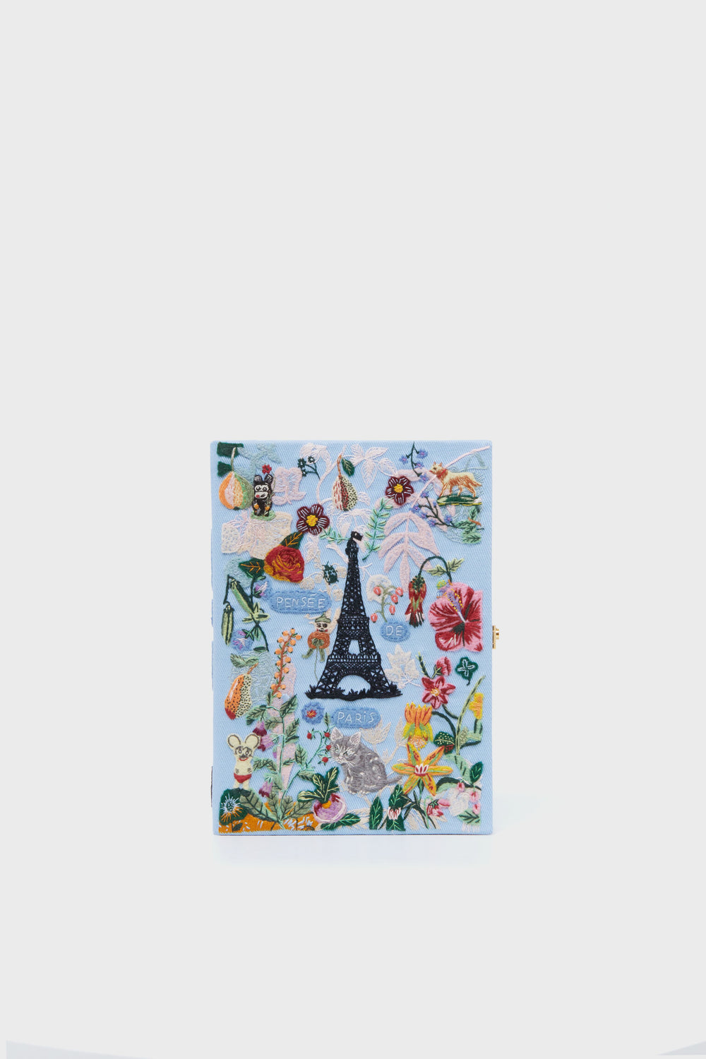 An Olympia Le-Tan light blue notebook stands upright, featuring colorful embroidered flowers, insects, and the Eiffel Tower on the cover, inspired by the brand's artisanal book clutch accessories.