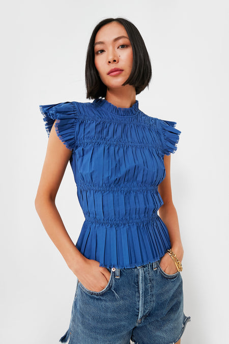 A woman in a pleated, short-sleeve cobalt Blue Greir Top by Sea New York, featuring ruffle details, styled with casual blue denim shorts.