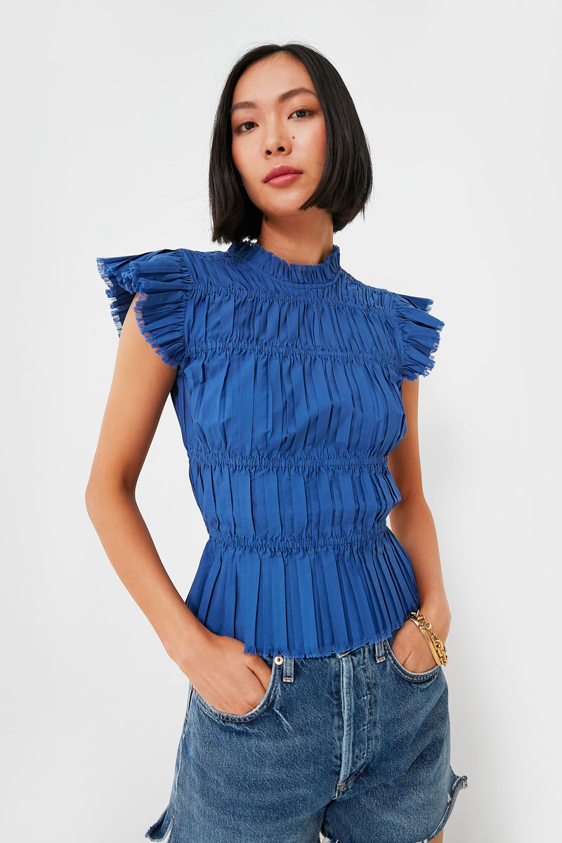 A woman in a pleated, short-sleeve cobalt Blue Greir Top by Sea New York, featuring ruffle details, styled with casual blue denim shorts.