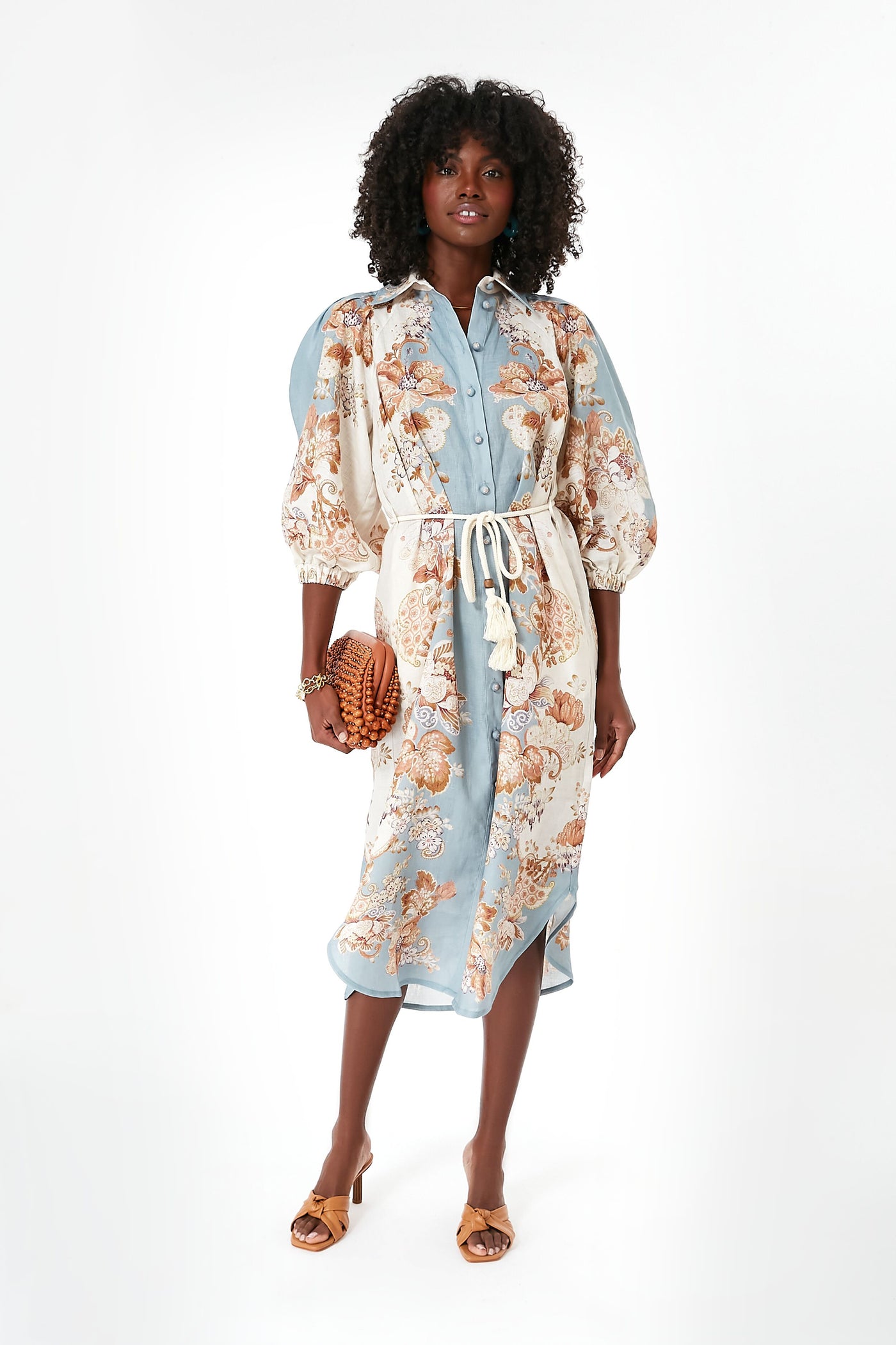 A woman wears an Alemais light blue and cream floral midi dress with puff sleeves and a tie belt, paired with brown heeled sandals and a woven clutch—perfect as a vacation dress.
