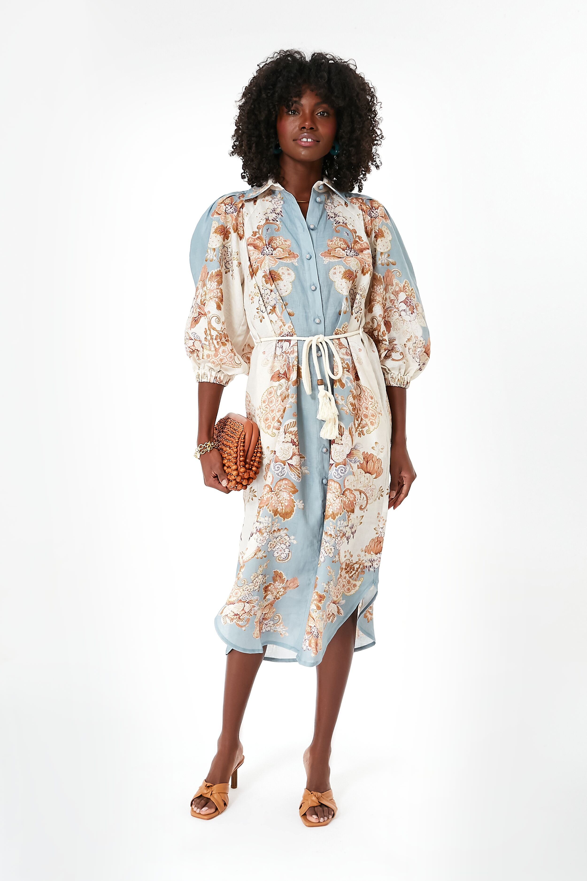 A woman wears an Alemais light blue and cream floral midi dress with puff sleeves and a tie belt, paired with brown heeled sandals and a woven clutch—perfect as a vacation dress.