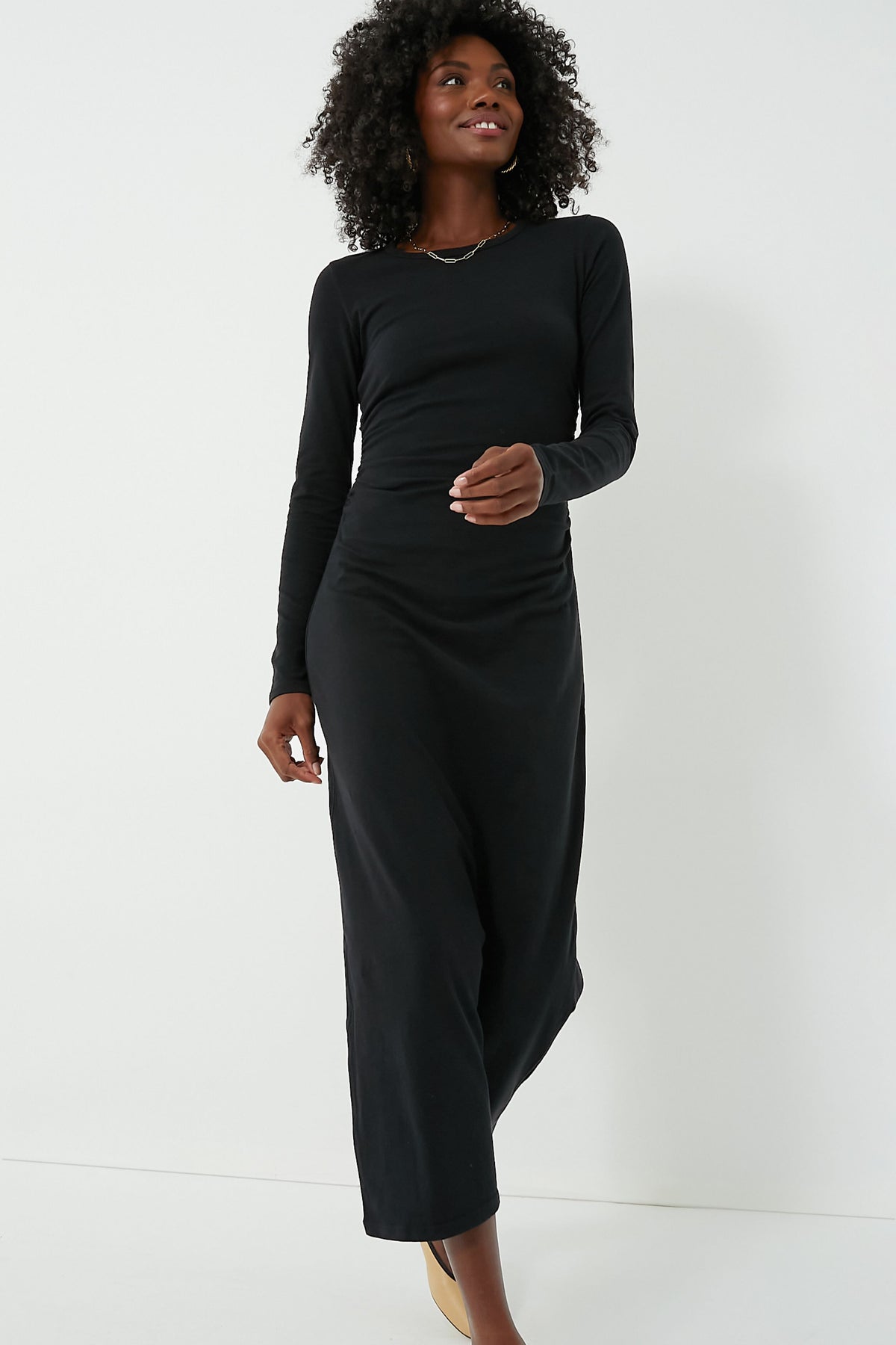 A person stands confidently wearing a long-sleeved Black Wiley Dress by XiRENA, featuring an elastic ruched waist, paired with light-colored shoes.