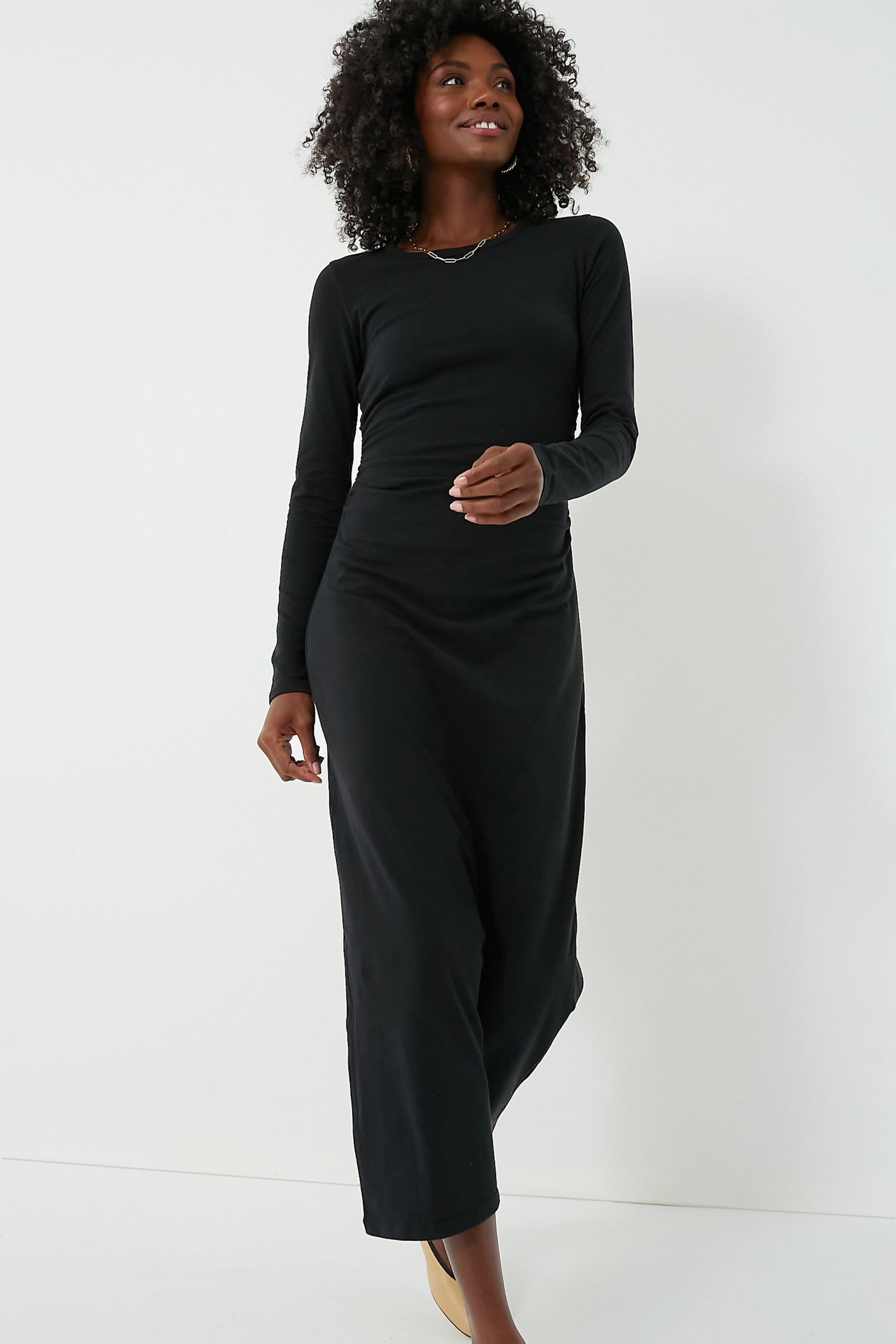 A person stands confidently wearing a long-sleeved Black Wiley Dress by XiRENA, featuring an elastic ruched waist, paired with light-colored shoes.