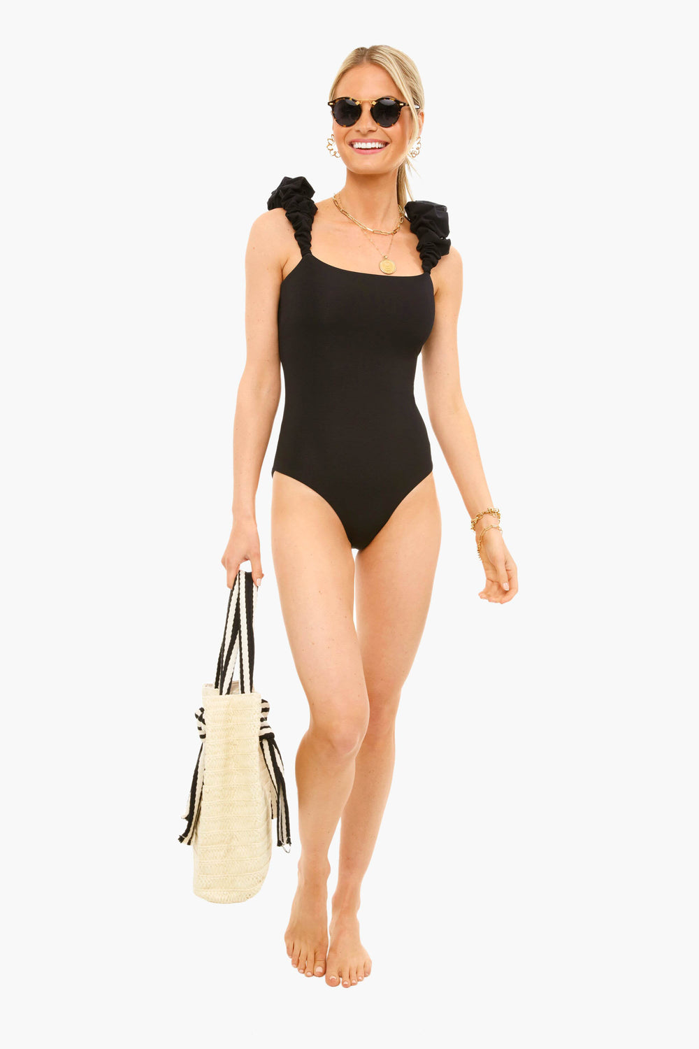A woman stands barefoot, wearing a Maygel Coronel black one-piece swimsuit with ruffle shoulders, accessorized with sunglasses, necklaces, bracelets, and holding a woven beach bag.