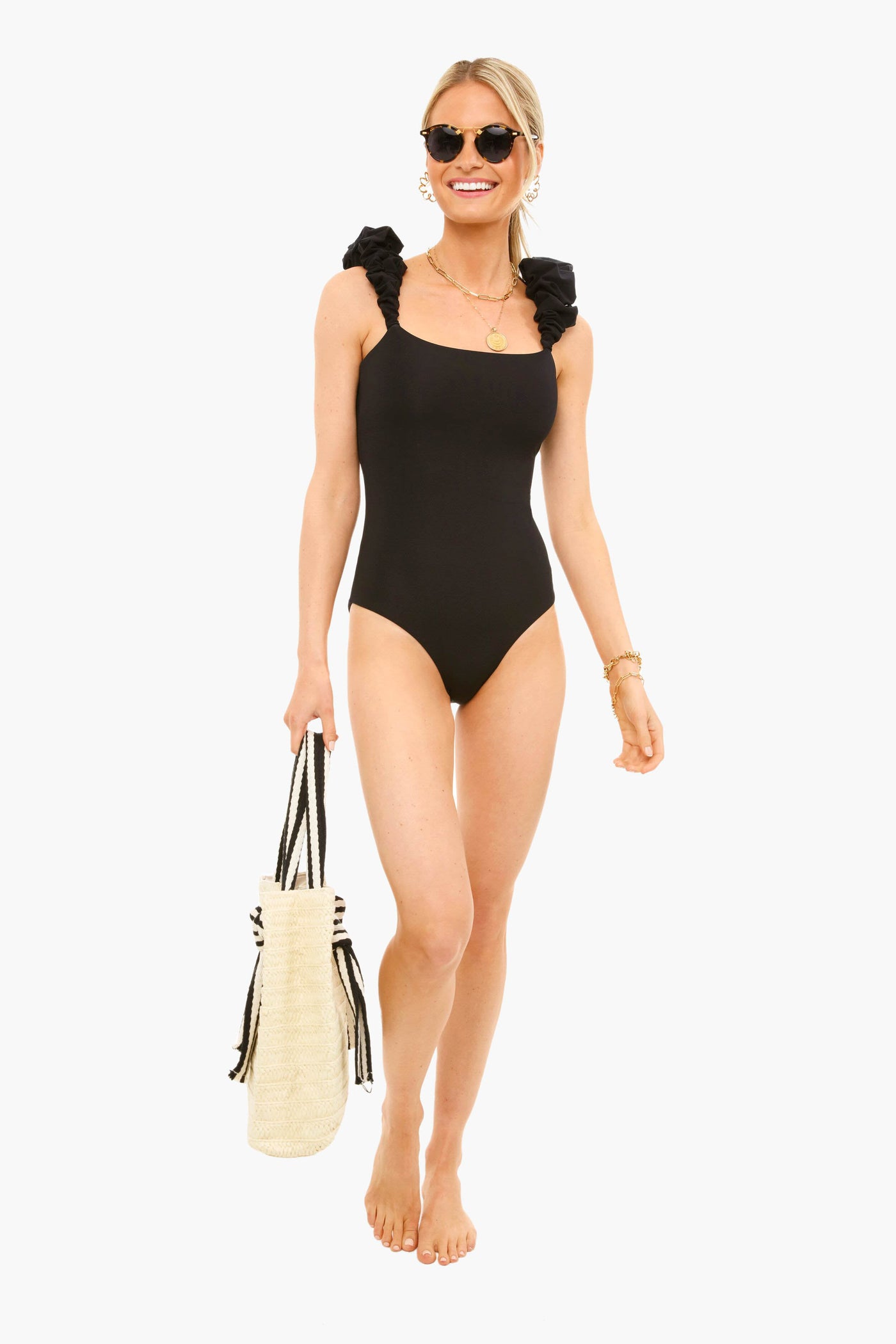 A woman stands barefoot, wearing a Maygel Coronel black one-piece swimsuit with ruffle shoulders, accessorized with sunglasses, necklaces, bracelets, and holding a woven beach bag.