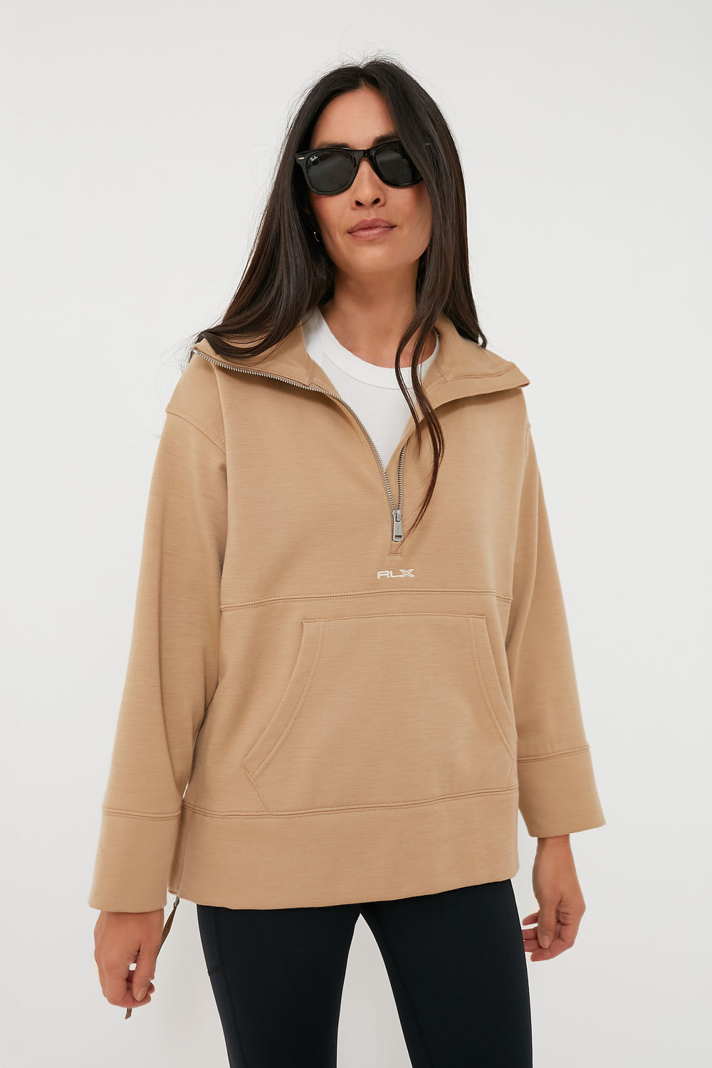 A woman wears RLX Ralph Lauren sunglasses, a beige wool blend quarter-zip pullover, a white shirt, and black leggings, confidently showcasing stylish golf apparel.