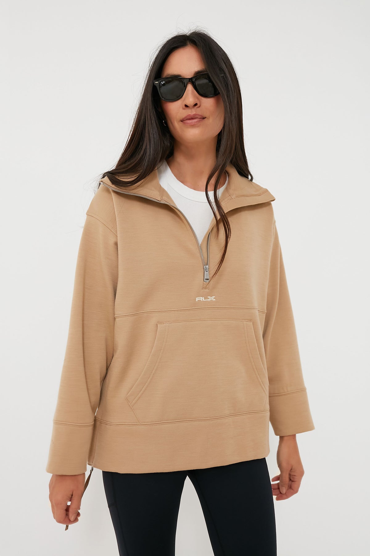 A woman wears RLX Ralph Lauren sunglasses, a beige wool blend quarter-zip pullover, a white shirt, and black leggings, confidently showcasing stylish golf apparel.