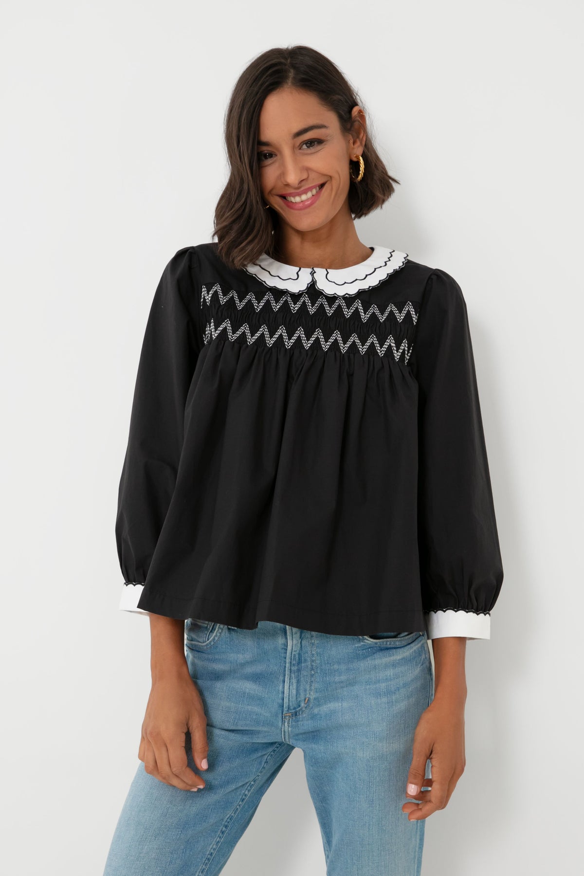 A woman wears a BATSHEVA Victorian-inspired black smocked blouse with a white scalloped collar and cuffs, styled with light blue jeans.