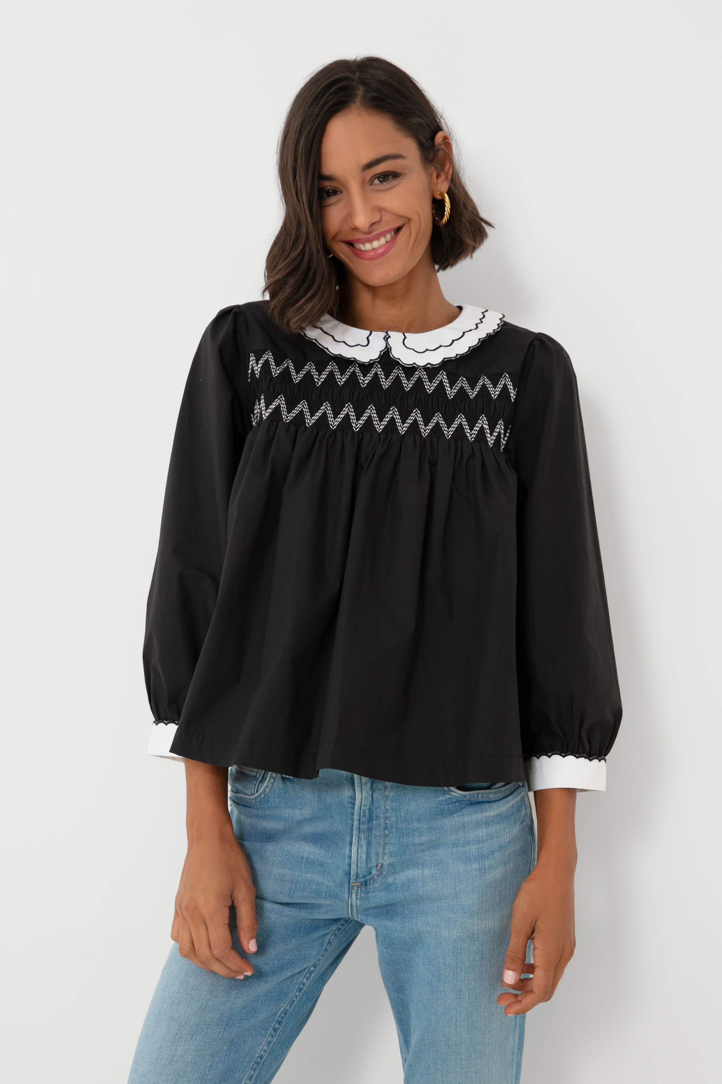 A woman wears a BATSHEVA Victorian-inspired black smocked blouse with a white scalloped collar and cuffs, styled with light blue jeans.