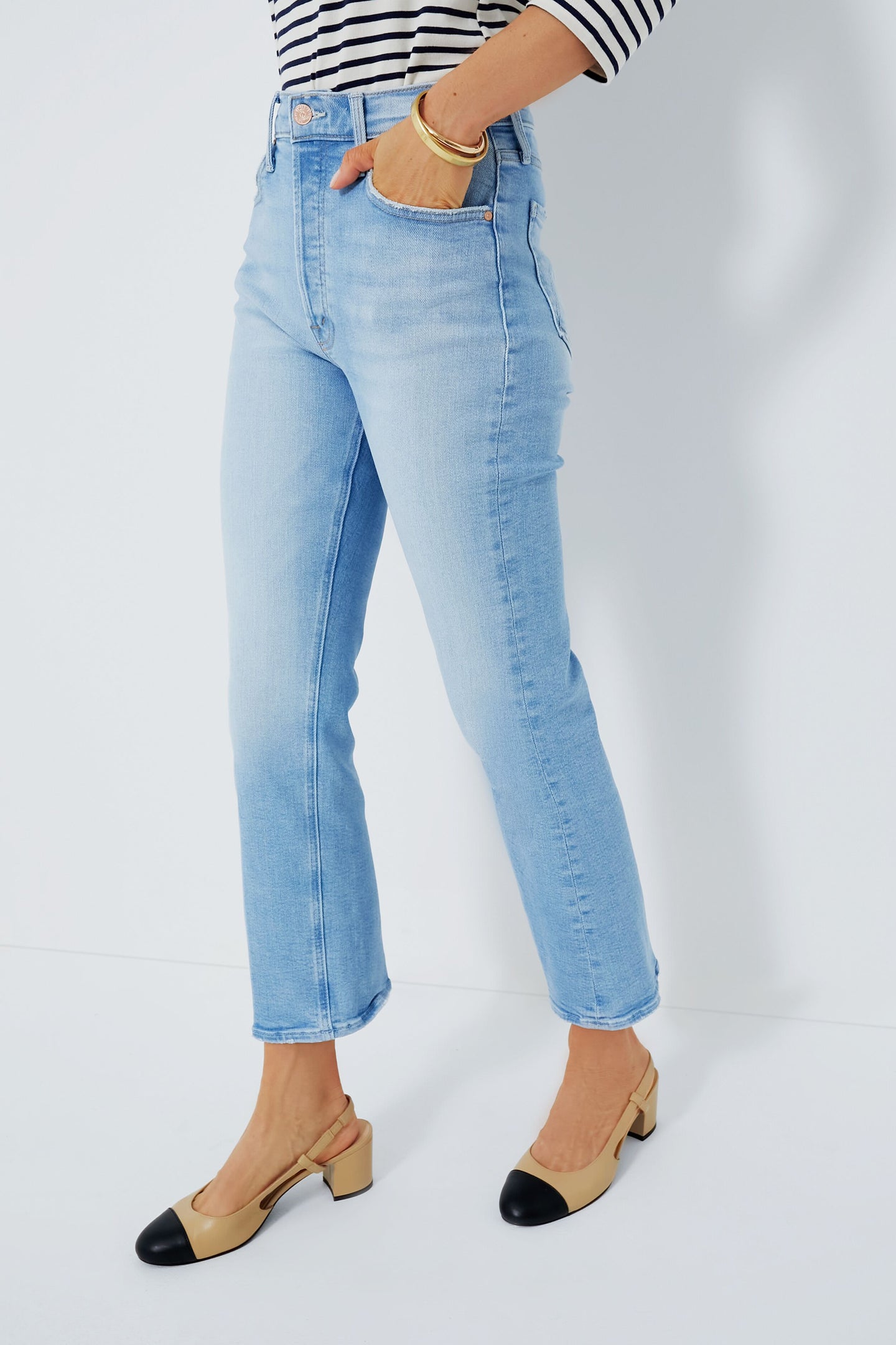 A person wears MOTHER high-rise ankle jeans in light blue, a striped long-sleeve shirt, and beige low-heeled shoes with black toe caps.