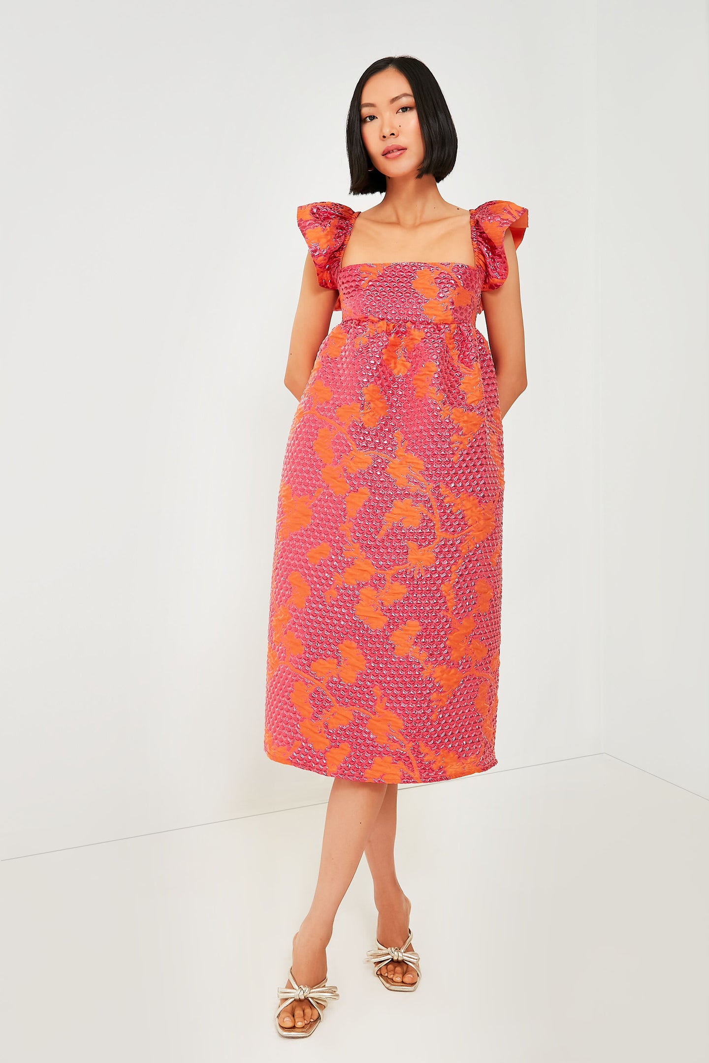 A woman wears a Baum Und Pferdgarten Pink Bubble Astraea Dress with ruffle sleeves and floral texture, standing in a minimalist white room with sandals—ideal for stylish events.