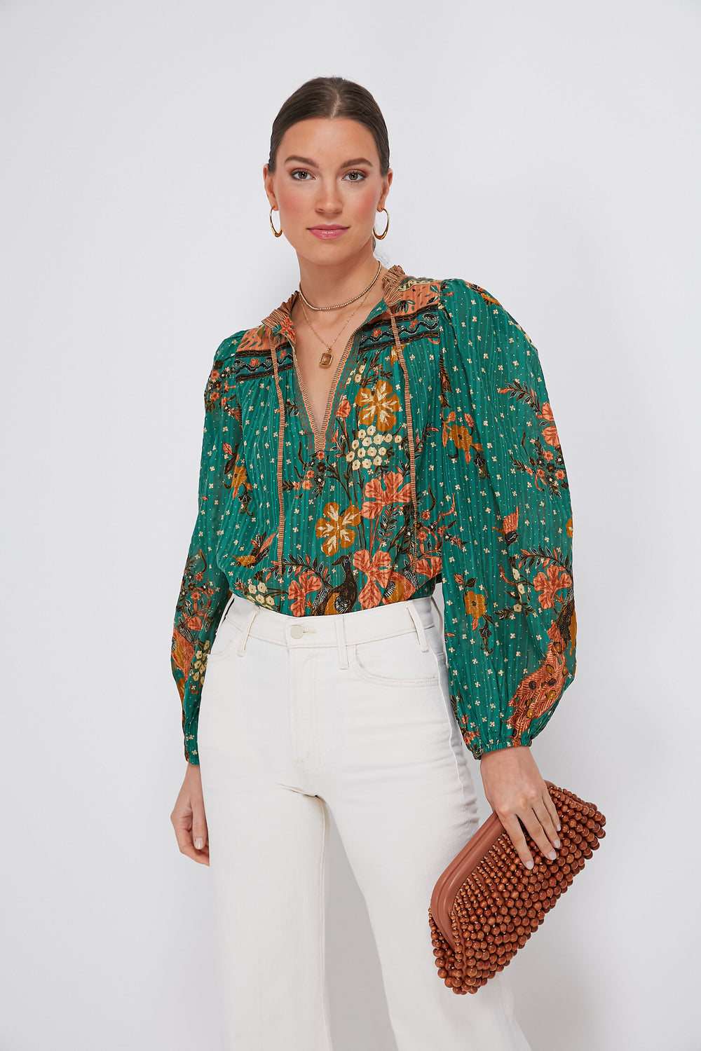 A woman in a green floral Ulla Johnson blouse and white high-waisted pants holds a brown textured clutch.