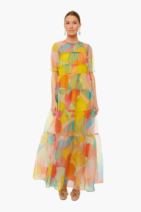 A woman wears the STAUD sheer organza maxi dress with abstract kaleidoscope print, short sleeves, and tiered layers. Her hair is tied back, and she accessorizes with earrings and sandals.