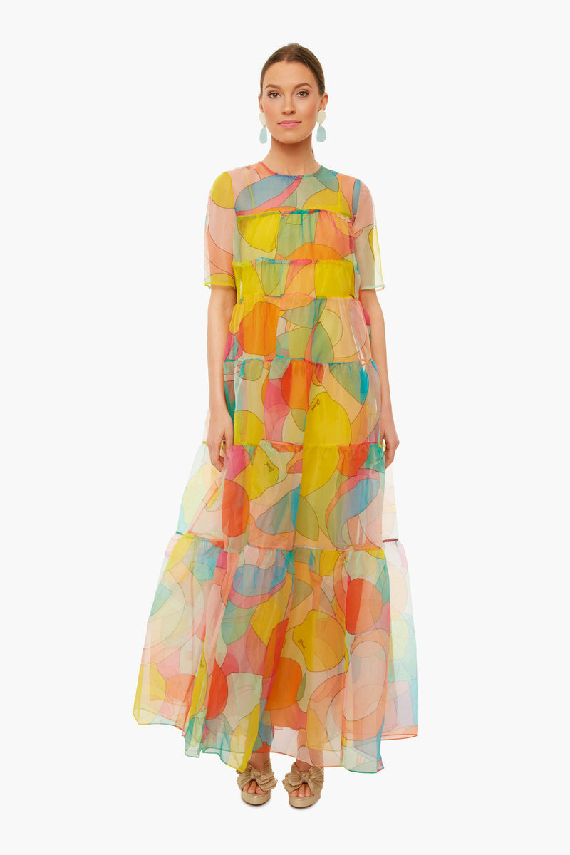 A woman wears the STAUD sheer organza maxi dress with abstract kaleidoscope print, short sleeves, and tiered layers. Her hair is tied back, and she accessorizes with earrings and sandals.