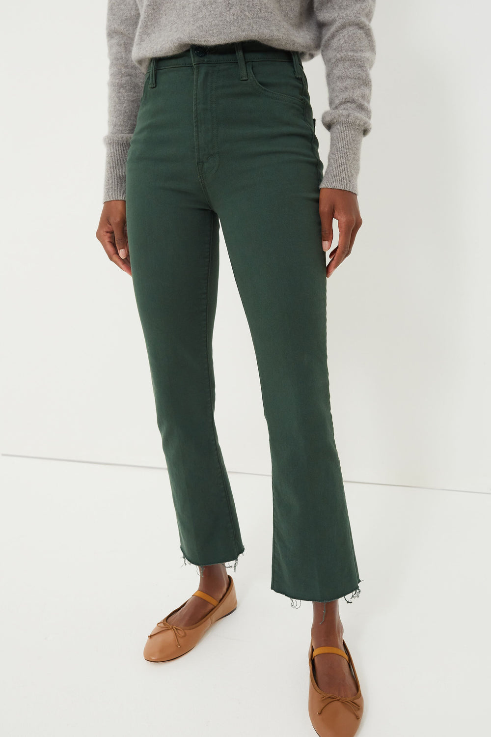 Person wearing a gray sweater, high rise ankle length green kick flare jeans with frayed hems from MOTHER, and tan ballet flats.