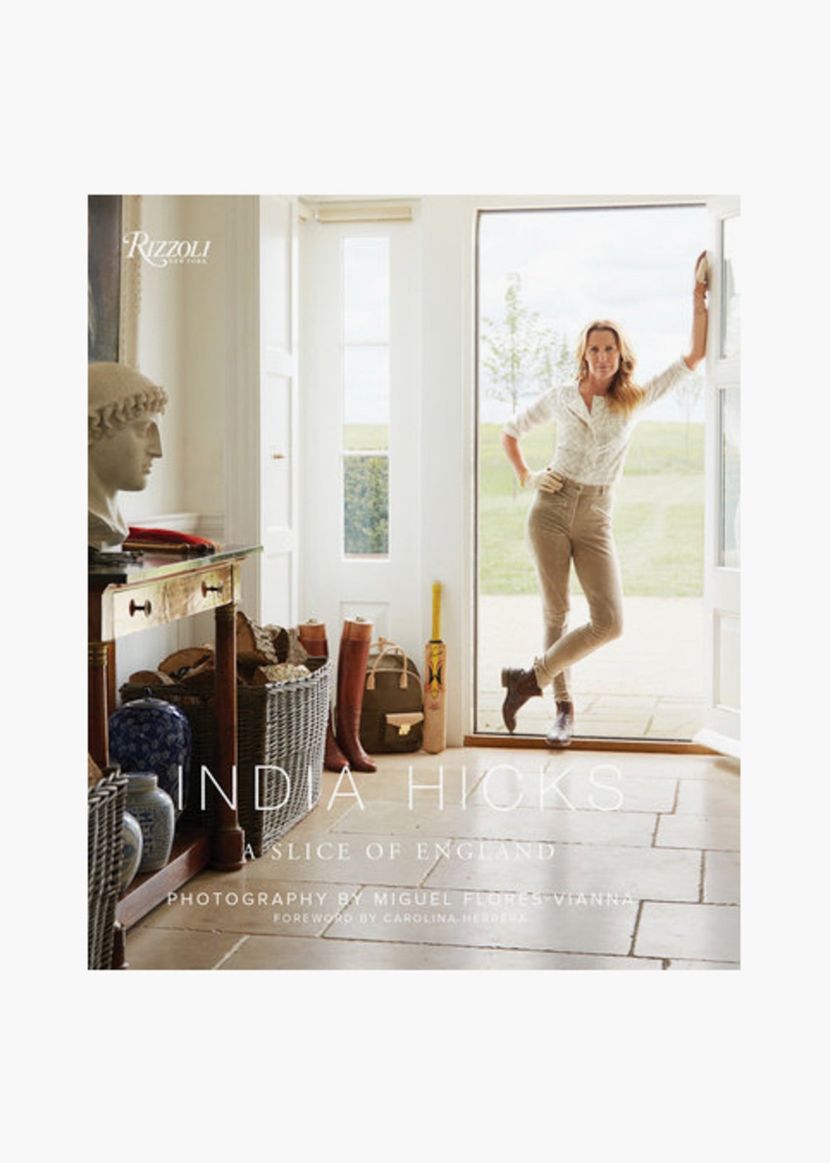 A woman stands in the doorway of a bright, elegant room with country decor. She holds "India Hicks: A Slice of England," a Rizzoli book showcasing the charm of English countryside designer homes.