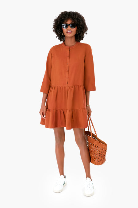A woman in an orange tiered Apiece Apart skirt dress pairs it with white sneakers, sunglasses, and a woven tote bag.