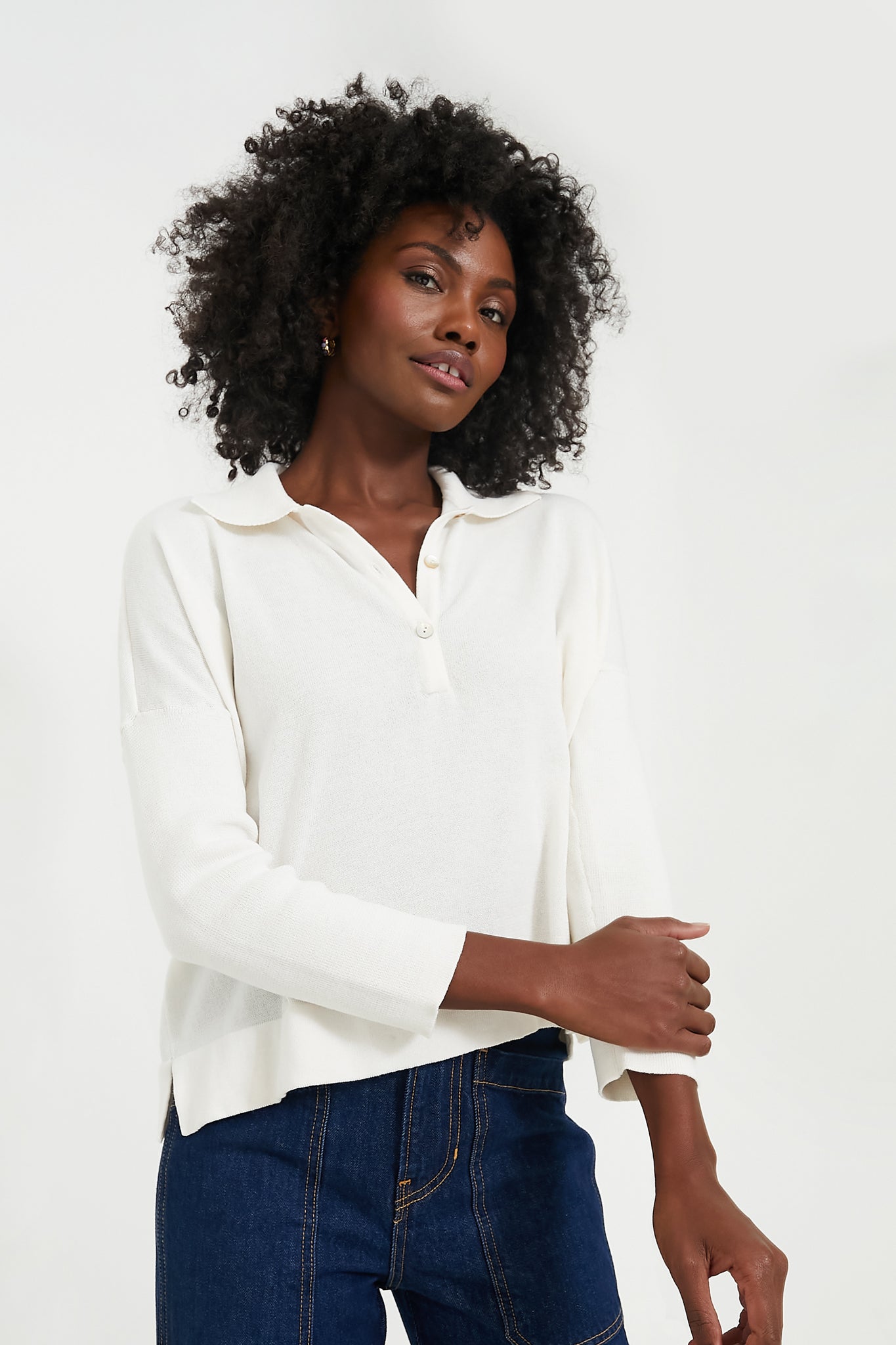 A woman with curly hair stands confidently, wearing a Tuckernuck long-sleeve white spread collar shirt with a partial button placket and dark blue jeans.