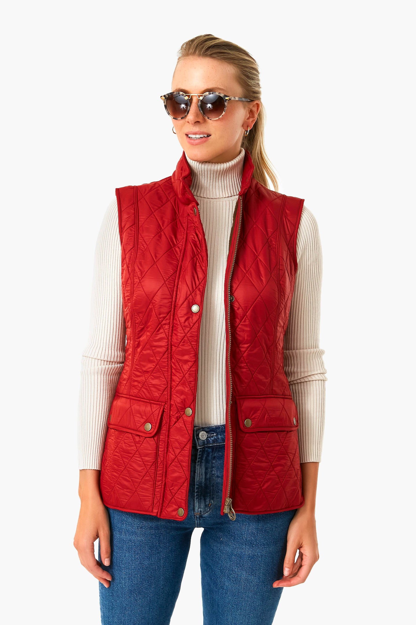 A woman wears a Barbour red quilted vest, styled like the Wray Gilet, layered over a white ribbed turtleneck, with blue jeans and sunglasses.