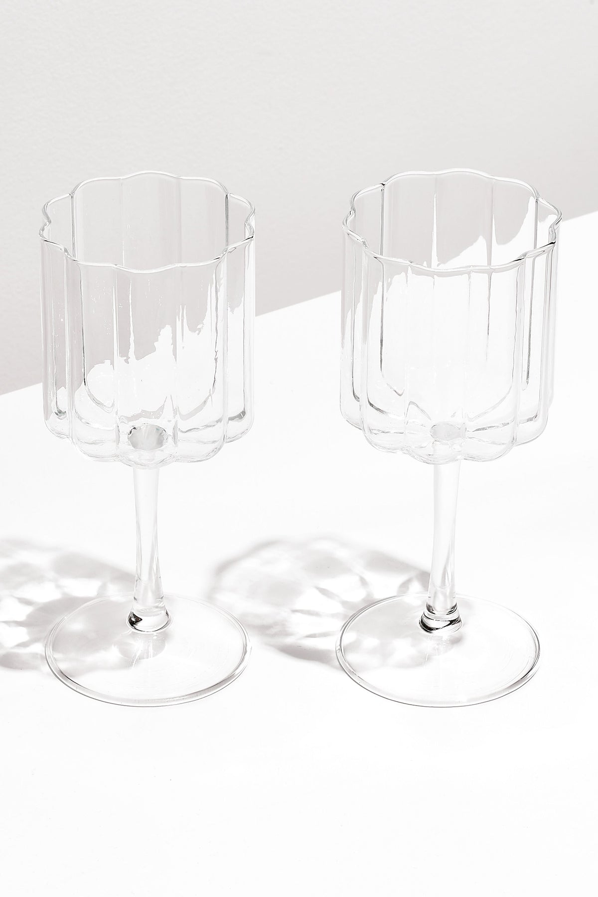 Two clear, scalloped-edge wave wine glasses by FAZEEK sit side by side on a white surface, crafted from hand-blown borosilicate glass for a sculptural homeware touch.
