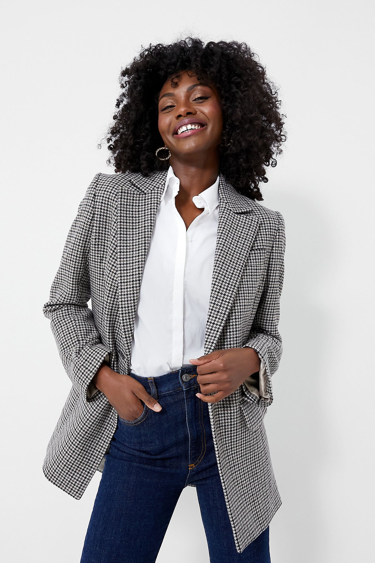 A smiling person with curly hair wears a Barbour micro-houndstooth blazer, white shirt, and blue jeans.