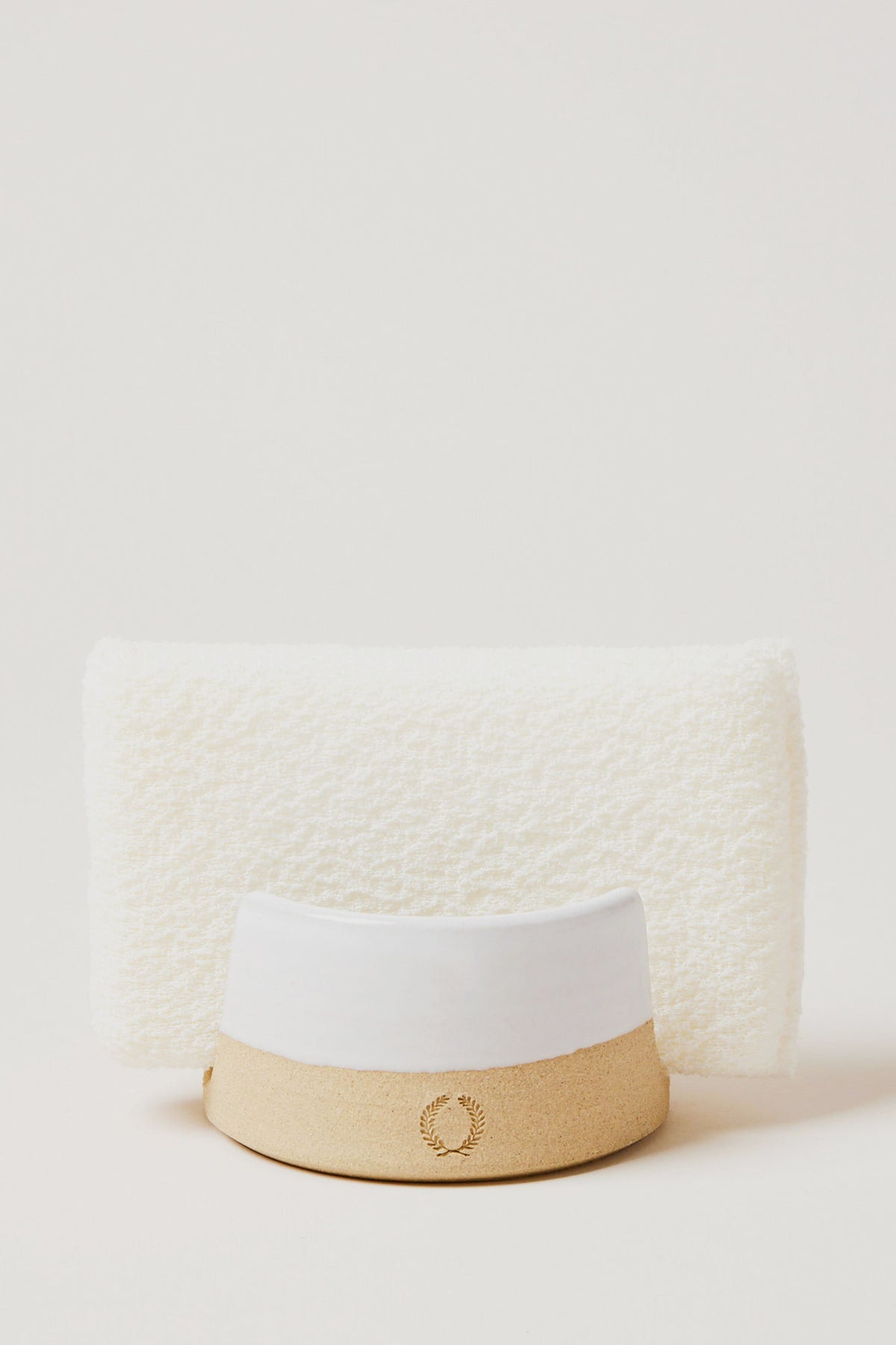 A white textured towel stands upright behind a beige and white hat with a laurel wreath emblem, alongside Farmhouse Pottery, Inc accents and a kitchen sponge holder.