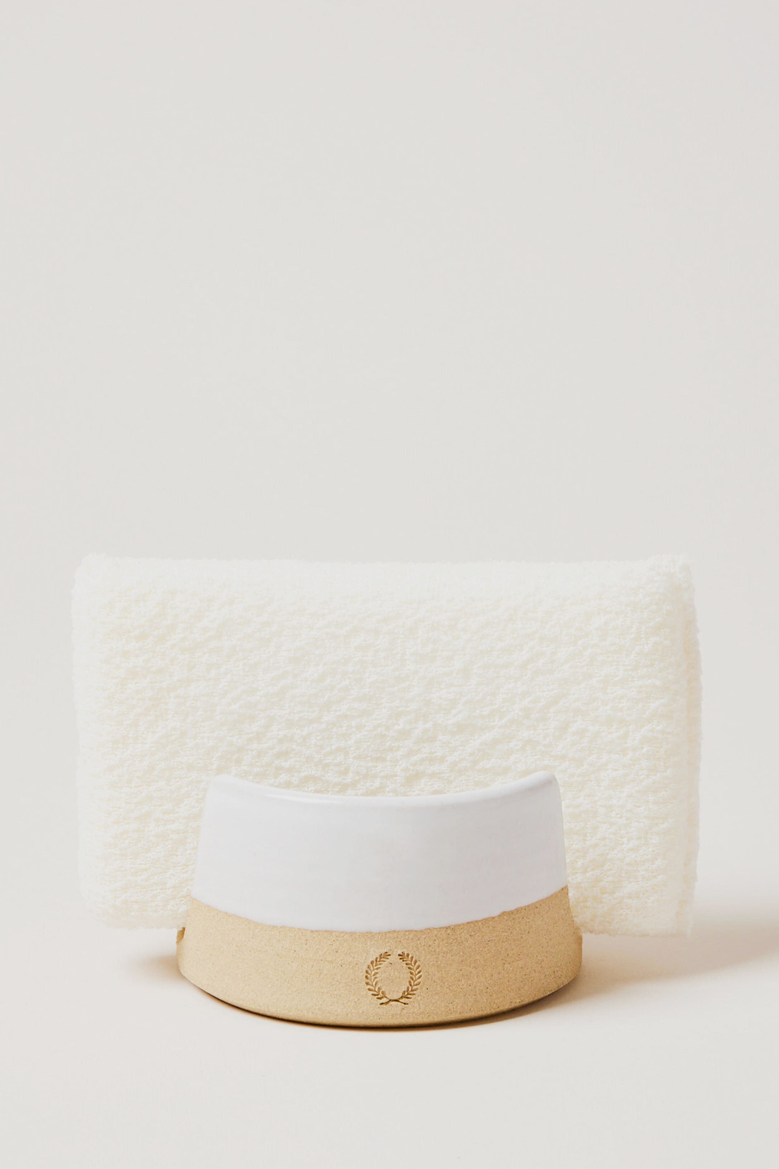 A white textured towel stands upright behind a beige and white hat with a laurel wreath emblem, alongside Farmhouse Pottery, Inc accents and a kitchen sponge holder.