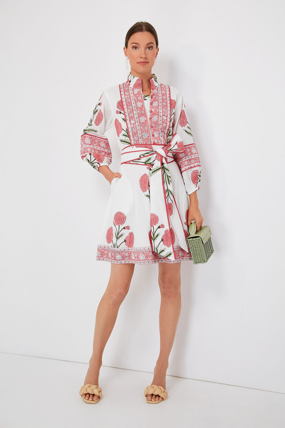 A woman wears the Sue Sartor Rosy Marigold Flounce Shorty Dress, featuring pink floral patterns and a matching waist tie, paired with a green woven handbag and beige sandals—crafted from sustainable fabrics for a modern look.