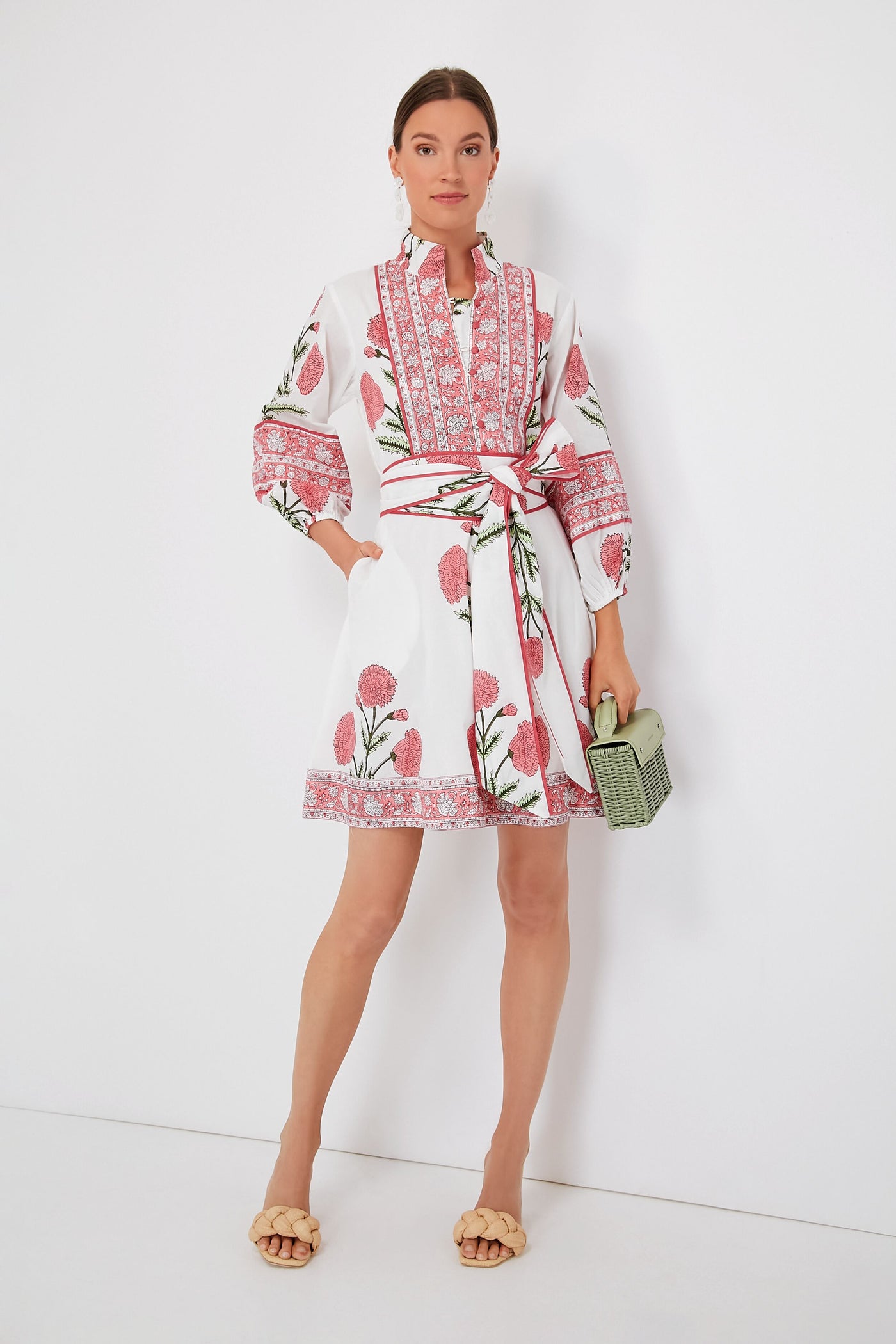 A woman wears the Sue Sartor Rosy Marigold Flounce Shorty Dress, featuring pink floral patterns and a matching waist tie, paired with a green woven handbag and beige sandals—crafted from sustainable fabrics for a modern look.